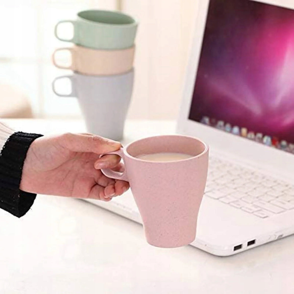 Dłoń trzyma różowy kubek IMODE przy otwartym laptopie; w tle stoją kubki w innych kolorach.